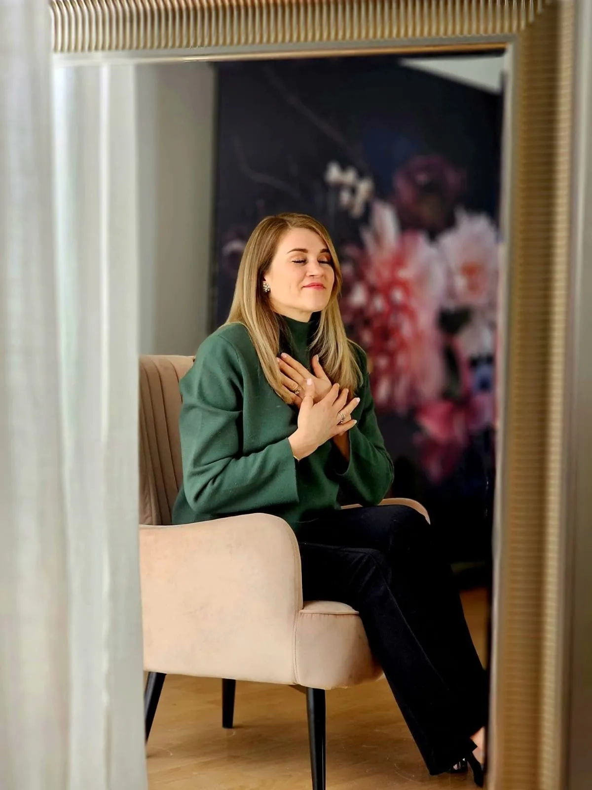 Natalie Leer im Sessel vor einer dunklen Blumentapete, eine Hand auf der Brust